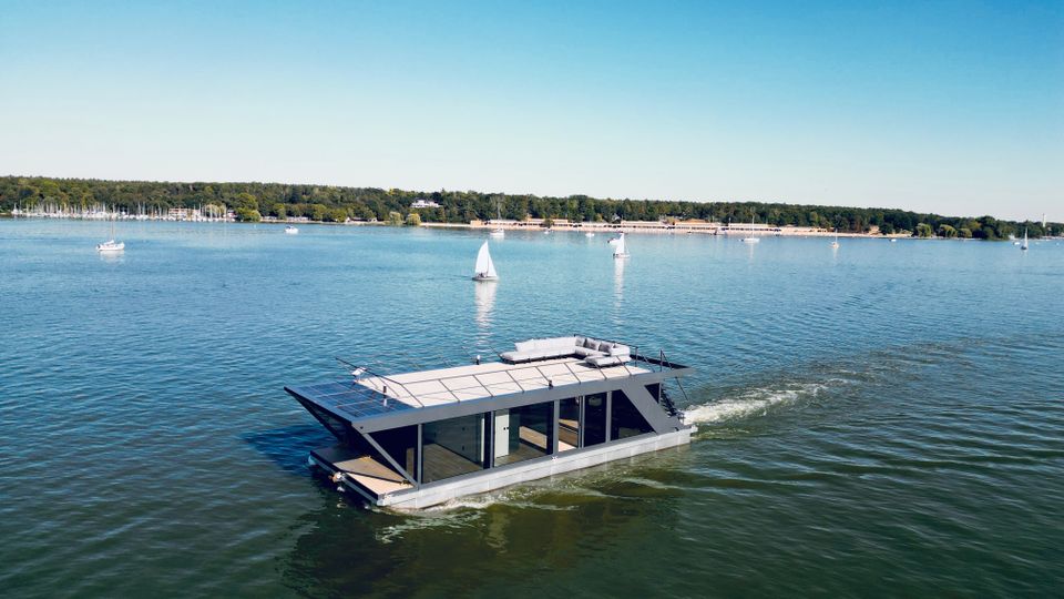 Ein modernes Hausboot mit einer großen Terrasse fährt über einen ruhigen See, umgeben von kleineren Segelbooten und einem Ufer mit Bäumen und Anlegestellen im Hintergrund.