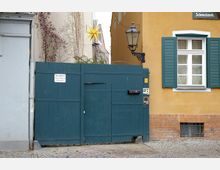 Eine dunkelgrüne Holztor-Einfahrt zwischen zwei Gebäuden, mit einem gelben Stern über der Mauer und einem Wandlaternenlicht. Rechts daneben befindet sich eine beige Wand mit einem Fenster und grünen Fensterläden, sowie einem Straßenschild mit der Hausnummer 37.