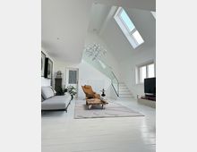 Modernes Wohnzimmer mit weiß gestrichenen Holzböden, hellem Sofa, braunem Designersessel und einem niedrigen Couchtisch auf einem Teppich. Eine Treppe mit Glasgeländer führt zu einem Obergeschoss, und Oberlichter sowie große Fenster lassen viel Tageslicht einfallen.