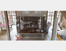 Eine moderne Loft-Innenansicht mit einer zentralen schwarzen Küche, flankiert von hohen Fenstern mit Sprossenrahmen. Im Hintergrund sind ein Billardtisch, eine Motorrad-Dekoration und eine Sitzecke sichtbar, die den industriellen Stil unterstreichen.