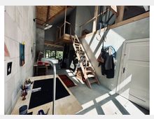 Blick von oben in einen hohen, hellen Innenraum mit Sichtbetonwänden, großen Fenstern und eingezogenem Holz-Zwischengeschoss mit Treppe und Geländer. Unten sind ein Fliesenboden sowie eine Werkbank mit Werkzeugen und Materialien zu sehen, durch die Fenster fällt starkes Sonnenlicht mit Schattenmustern.