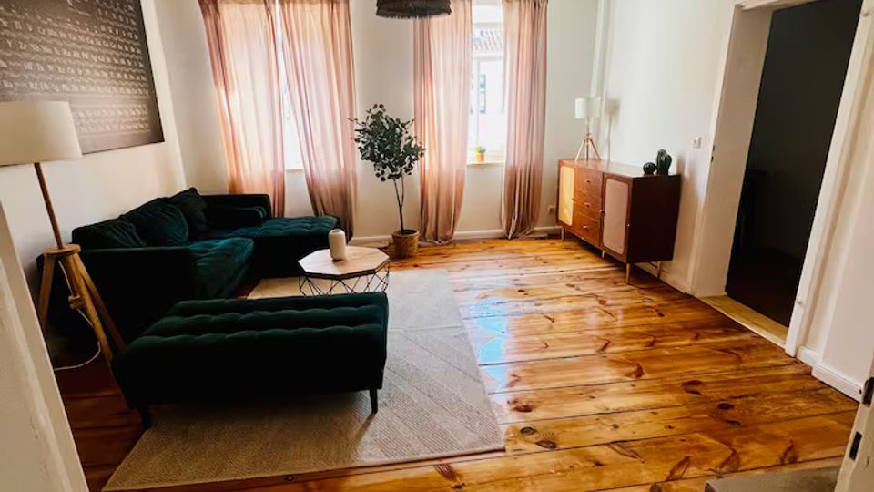 Helles Wohnzimmer mit breiten Holzdielen und sichtbaren Deckenbalken, zwei Fenstern mit hellen Vorhängen und einer dunklen Pendelleuchte. Dunkelgrünes Sofa mit Couchtisch auf einem Teppich sowie eine Kommode an der rechten Wand.