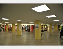 Unterirdische Passage mit gelb gefliesten Säulen, gleichmäßig beleuchtet durch rechteckige Deckenlampen. An den Seiten befinden sich kleine Geschäfte, darunter Bäckereien und ein Verkaufsstand mit Obst und Blumen.