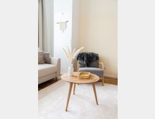 Helles Wohnzimmer mit einem runden Holztisch, auf dem sich eine weiße Vase mit getrocknetem Pampasgras und ein kleiner Korb befinden. Im Hintergrund stehen ein grauer Sessel mit einer dunklen Decke darüber und ein beigefarbenes Sofa auf einem hellen Teppich.