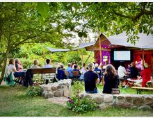Eine Outdoor-Veranstaltung in einem Garten mit Menschen, die auf Holz- und Klappstühlen sitzen, unter einem weißen Sonnensegel im Schatten von Bäumen. Im Hintergrund eine Steinmauer und eine Leinwand mit einer Präsentation.