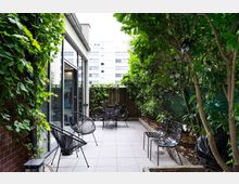 Small urban patio beside a modern building with large glass doors and light gray tile flooring. The narrow courtyard is enclosed by dense trees and climbing vines, with black metal chairs and a small table, bicycles near a wooden fence, and string lights hanging in the greenery.