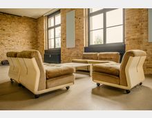 Loftartiger Innenraum mit unverputzten Ziegelwänden, großen Sprossenfenstern und Heizkörpern unter den Fenstern. Im Vordergrund stehen beige Polstersofas auf Rollen um einen niedrigen weißen Couchtisch auf glattem Betonboden.