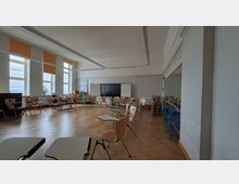 Heller Klassenraum mit großen Fenstern und orangefarbenen Rollos, Parkettboden und in einem Halbkreis angeordneten Stühlen mit Schreibflächen. Vorn stehen ein großer Bildschirm zwischen Whiteboards sowie Regale und Materialwagen an der Wand.