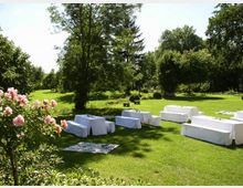 Grüne Parklandschaft mit mehreren weiß bedeckten Sitzbänken, die auf einer Rasenfläche angeordnet sind, umgeben von Bäumen und Blumen, darunter blühende Rosen im Vordergrund. Der Bereich vermittelt den Eindruck einer Veranstaltung im Freien.