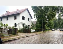 Wohnhaus mit weißer Fassade und rotem Ziegeldach, umgeben von einem kleinen Garten mit Bäumen und Pflanzen. Im Vordergrund eine Kopfsteinpflasterstraße mit parkenden Autos, gesäumt von großen Bäumen.