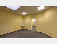 Leerer Innenraum mit gelben Wänden, grauem Teppichboden und einer abgehängten Rasterdecke mit integriertem Beleuchtungssystem. An der linken Wand ist ein kleines Waschbecken montiert, rechts führt eine Tür in einen weiteren Raum mit vertikalen Jalousien.