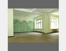 Ein leerer Raum mit hellen Wänden, Holzfußboden und großen Fenstern, die Tageslicht hereinlassen. Eine Wand ist mit einem dekorativen grünen Muster gestaltet, das an geschwungene Bögen erinnert.