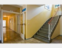 Ein heller Innenflur mit einer Treppe aus grauem Stein und einem roten Handlauf auf der rechten Seite. Links führen Türen zu einem beleuchteten Korridor, und ein Schild mit "WC" weist auf die Toiletten hin.