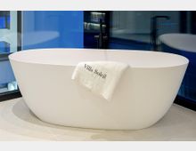 Moderne Badezimmeransicht mit einer freistehenden, ovalen weißen Badewanne auf hellen Fliesen. Dahinter sind eine schwarze Standarmatur und große Glasflächen mit blau getönten Spiegelungen zu sehen.