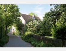 Pflanzenreicher Weg mit einem kleinen, gemauerten Haus mit Ziegeldach im Hintergrund. Umgeben von blühenden Sträuchern, Bäumen und einem Holzzaun; leicht schattige Atmosphäre unter einem klaren Himmel.