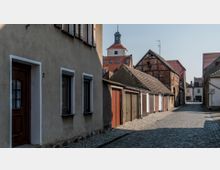 Eine gepflasterte Gasse in einer historischen Kleinstadt mit einfachen Häusern, Garagen und Fachwerkgebäuden. Im Hintergrund ist ein Kirchturm mit Uhr und rotem Dach zu sehen.
