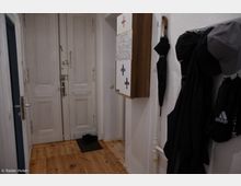 Flur mit holzfarbenem Laminatboden, einer weißen Eingangstür mit Briefschlitz und mehreren Sicherheitsriegeln. Rechts an der Wand befinden sich eine Garderobe mit Kleidungsstücken, Hüten und ein Regenschirm; darüber hängt ein Schrank mit Aufklebern und einer Karte.