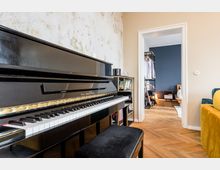 Innenraum einer Wohnung mit schwarzem Klavier im Vordergrund vor einer hellen, teils abgenutzten Wand und Fischgrätparkettboden. Durch einen weißen Türrahmen blickt man in ein angrenzendes Zimmer mit dunkelblauer Wand, Kleiderständer und niedrigem Bett.