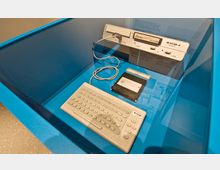 Historischer Heimcomputer KC 85/4 aus der DDR, ausgestellt in einer Glasvitrine. Zu sehen sind die Basiseinheit, eine angeschlossene Tastatur, ein Datenkabel und eine Diskette.