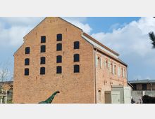 Backsteinbau mit Satteldach und mehrfachen, dunkel verglasten Fenstern sowie grünen Fensterläden auf der rechten Gebäudeseite. Im Vordergrund befindet sich eine Grünfläche mit einer Statue, im Hintergrund ein weiteres Gebäude aus Ziegelstein und ein blauer Himmel mit Wolken.