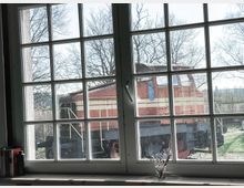 Blick durch ein mehrteiliges, weiß gerahmtes Fenster auf eine rote Rangierlokomotive mit beiger Streifenlackierung, die auf einem Außengelände steht. Im Vordergrund auf der Fensterbank befinden sich ein kleines Blumenarrangement und Bücher.