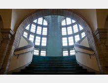 Großer, geschwungener Treppenaufgang in einem historischen Gebäude, eingerahmt von einem steinernen Rundbogen. Im Hintergrund sind hohe, halbkreisförmige Fenster und eine hellblaue Wand mit dekorativen Mustern sichtbar.