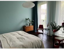 Helles Schlafzimmer mit mintfarbener Wand, großem Fenster mit transparenten Vorhängen und dunklem Holzboden. Neben dem Bett stehen eine Holzkommode mit Pflanze, ein kleiner runder Beistelltisch und rote Stühle an einem Glastisch.