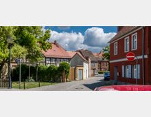 Straßenszene in einem Dorf mit Kopfsteinpflaster, umgeben von Fachwerkhäusern, roten Ziegeldächern und einem roten Gebäude mit weißen Fensterläden im Vordergrund. Links steht eine Straßenlaterne, und am Straßenrand ist ein "Einfahrt verboten"-Schild sichtbar.