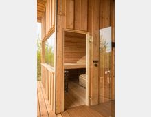Eine Holzhütte mit einer offenen Tür, die den Blick in einen kleinen Sauna-Innenraum mit Holzbänken und einem Saunaofen freigibt; außen und innen bestehen die Wände aus unbehandelten Holzbrettern. Im Hintergrund sind Bäume durch eine Glasfläche sichtbar.