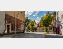 Straßenszene in einem Wohngebiet mit Kopfsteinpflaster, alten Gebäuden und einer gelb gestrichenen Fassade im Hintergrund. Ein Baum steht zentral neben einem Verkehrsschild, während links ein historisches Gebäude mit roten Ziegeln und rechts ein modernes Gebäude mit Glasdetails erkennbar ist.