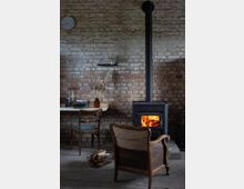 Ein rustikaler Raum mit unverputzter Ziegelwand, in dem ein Holzofen mit brennendem Feuer im Mittelpunkt steht. Im Vordergrund ein Holzsessel mit Stoffbezug, daneben ein Korb mit Feuerholz; links ein Schreibtisch mit Schreibmaschine und Dekoration.