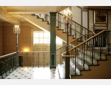 Treppenhaus in einem historischen Gebäude mit eleganten Metallgeländern, Marmorstufen und dekorativen Säulen. Im Hintergrund befinden sich große Fenster, ein Doppeltür-Eingang sowie ein Hinweisschild zum "Großen Saal" und "Kleinen Saal".