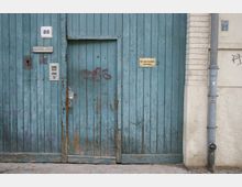 Alte, blau gestrichene Holztür mit sichtbaren Abnutzungsspuren, eingebaut in eine Mauer aus hellem Putz und Ziegeln. Neben der Tür befinden sich eine Hausnummer "88", eine Gegensprechanlage, ein Briefschlitz und ein Hinweisschild mit der Aufschrift "Ein- und Ausfahrt freihalten".