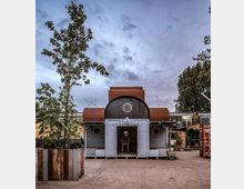 Kleines, holzverkleidetes Gebäude mit bogenförmigem Dach und runden Fenstern, umgeben von Bäumen, Pflanzenkübeln und einem offenen Platz. Im Hintergrund sind Container, ein Backsteingebäude und eine bewölkte Abendhimmelkulisse zu sehen.
