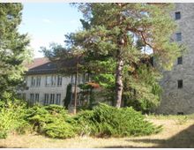 Außenansicht eines Gebäudekomplexes, bestehend aus einem verputzten Haus mit Satteldach und weißen Fenstern, sowie einer angrenzenden Mauer aus Natursteinen. Im Vordergrund befinden sich Bäume und dichte Sträucher auf einer grasbewachsenen Fläche.