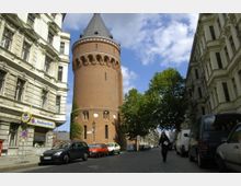 Straßenszene mit einem hohen, runden Backsteinturm mit Spitzdach in der Mitte, flankiert von historischen Wohngebäuden mit verzierten Fassaden. Geparkte Autos säumen die Kopfsteinpflasterstraße, während Bäume und blauer Himmel die Szenerie ergänzen.