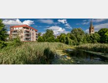 Wohngebäude mit moderner Fassade neben einem kleinen Teich und dichter Vegetation, im Hintergrund ein Kirchturm mit spitzer Dachkonstruktion. Der Himmel ist klar und von weißen Wolken durchzogen.