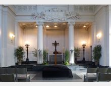 Innenraum einer kleinen Kapelle mit einem Altarbereich im klassizistischen Stil. Der Altar ist mit einem Holzkreuz, Kerzenhaltern und Pflanzen geschmückt, flankiert von zwei weißen Säulen und dekorativen Reliefs an der Wand.