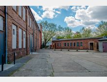 Ehemaliges Industriegebäude aus rotem Backstein mit hohen rechteckigen Fenstern und einer gepflasterten Freifläche, umgeben von Bäumen im Hintergrund unter einem teils bewölkten Himmel. Links ein zweistöckiges Gebäude und rechts ein einstöckiger Anbau.