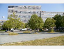 Eine städtische Szene mit einem großen, rechteckigen Wohn- oder Bürogebäude im Hintergrund, das von mehreren Etagen mit Fenstern geprägt ist. Im Vordergrund befinden sich eine Reihe von Bäumen, ein Platz mit Kiesboden und einige parkende Autos sowie ein Heißluftballon am Himmel.