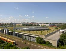 Außenansicht des Velodroms in Berlin, einem flachen, runden Gebäude mit begrüntem Dach, umgeben von einer offenen, parkähnlichen Fläche mit jungen Bäumen. Im Vordergrund sind Bahngleise und ein vorbeifahrender Zug sichtbar, im Hintergrund Wohngebäude und eine städtische Skyline unter klarem Himmel.
