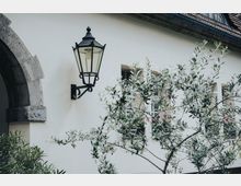 Außenansicht eines hell verputzten Gebäudes mit schwarzer, klassischer Wandlaterne und einem steinernen Rundbogen am linken Rand. Im Vordergrund stehen Zweige eines Baumes; dahinter sind Fenster mit Blumenkästen unter einem Dach mit Regenrinne zu sehen.