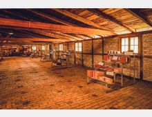 Innenraum einer historischen Ziegelfabrik mit freiliegenden Holzbalken und Ziegelboden. Am Boden stehen Schaukästen und Regale mit verschiedenen Ziegelmustern; die Wände sind aus Backstein und teils mit Fachwerk verstärkt, Licht fällt durch rechteckige Fenster ein.