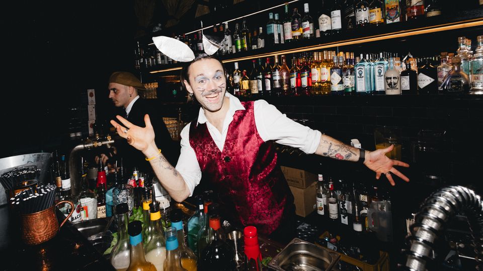 Innenansicht einer dunklen Cocktailbar mit schwarzer Fliesenwand, langen Regalreihen voller Spirituosenflaschen und warmen Lichtleisten. Im Vordergrund steht eine dicht bestückte Barstation mit Flaschen, Shakern, Eis und Barwerkzeugen.