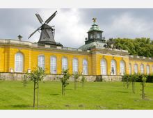 Gelbes Barockgebäude mit großen Sprossenfenstern, gekrönt von einer dekorativen Kuppel mit Uhr und einer Windmühle an der linken Seite; im Vordergrund ein gepflegter Rasen mit jungen Bäumen.