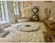 Heller Raum mit meditativer Einrichtung: ein großer, flauschiger Teppich bedeckt den Holzboden, darauf ein rundes Kissen mit verzierten Gongs im Hintergrund. Wände sind hell verputzt mit erdigen Farbakzenten, und es gibt helle Möbel und Kissen, die eine gemütliche Atmosphäre schaffen.