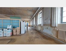 Ein langer, spärlich möblierter Raum mit hohen Fenstern auf der rechten Seite, teilweise mit Vorhängen verdeckt, und einer Trennwand aus Milchglas auf der linken Seite. Im Vordergrund stehen Kartons und Aktenboxen, während der Boden Gebrauchsspuren aufweist und die Wände teils beschädigt sind.