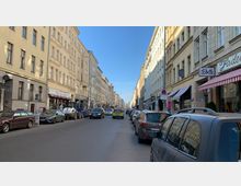 Straße in einer Stadt mit mehrstöckigen Wohn- und Geschäftshäusern aus hellem Putz und symmetrischen Fenstern. Geparkte Autos säumen beide Seiten der Straße, während sich auf der Fahrbahn mehrere Fahrzeuge bewegen; Geschäfte mit Schildern und Markisen befinden sich im Erdgeschoss der Gebäude.