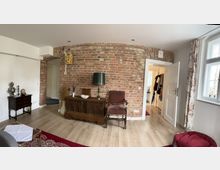 Wohnzimmer mit einer freiliegenden Backsteinwand, vor der historische Möbelstücke wie eine hölzerne Kommode, ein armlehnloser Sessel und eine Tischlampe stehen. Rechts befindet sich eine offene Tür, die in einen Flur führt, links ein kleines Sideboard; große Fenster bringen Tageslicht in den Raum.