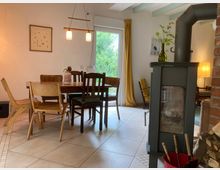 Gemütliches Wohnzimmer mit einem Holztisch, umgeben von verschiedenen Stühlen, und einem Kaminofen neben einem Stapel Holz. Im Hintergrund befinden sich eine große Glastür mit gelbem Vorhang, die ins Grüne führt, sowie eine Stehlampe und eine Sitzgelegenheit.